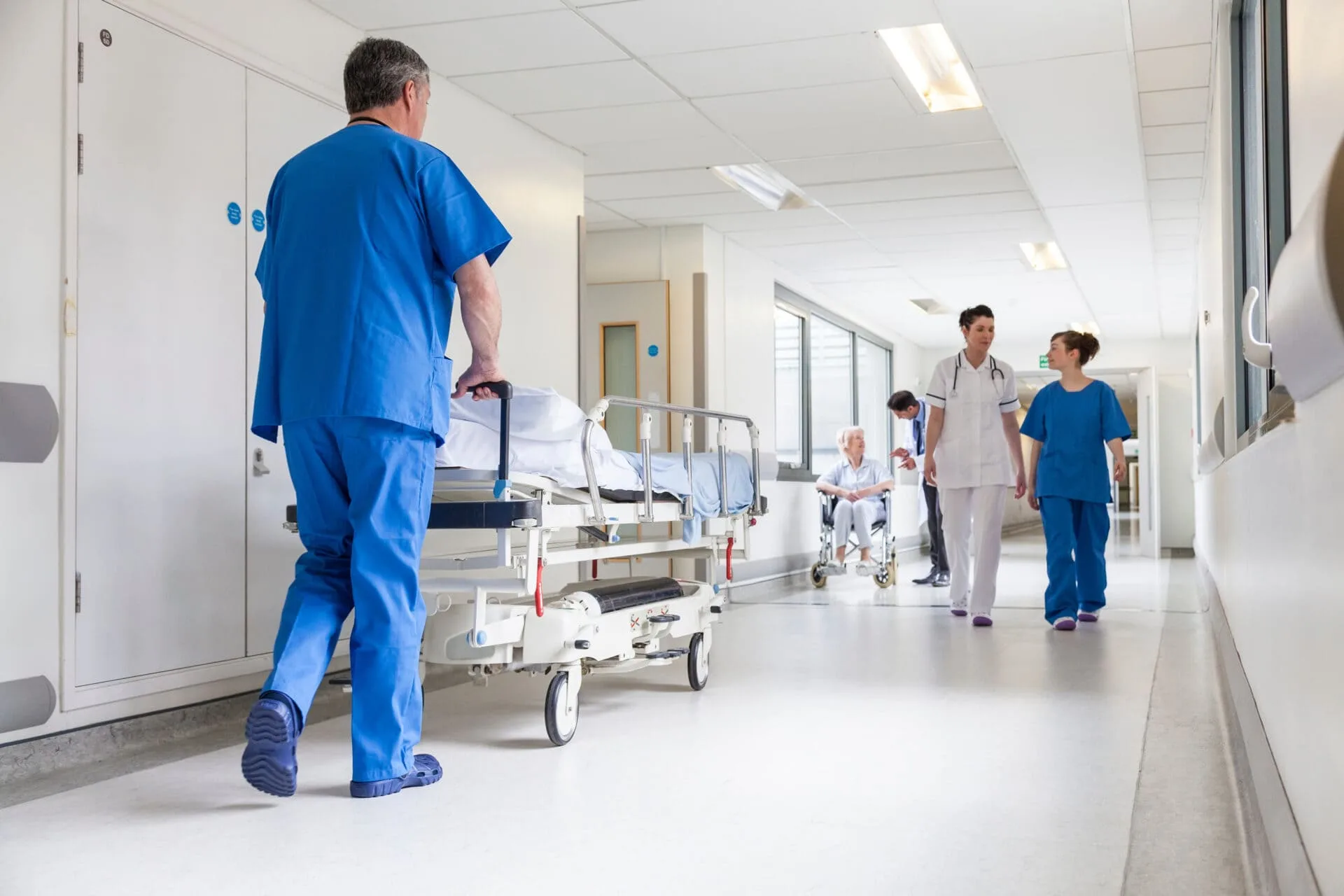 Male nurse pushing stretcher gurney bed in hospital corridor with doctors & senior female patient